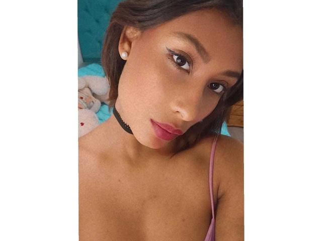 VanessaLove has brown skin, straight brown hair, and pink lipstick. She's wearing a black choker and pink spaghetti strap top, showing small tits. She's looking at the camera.