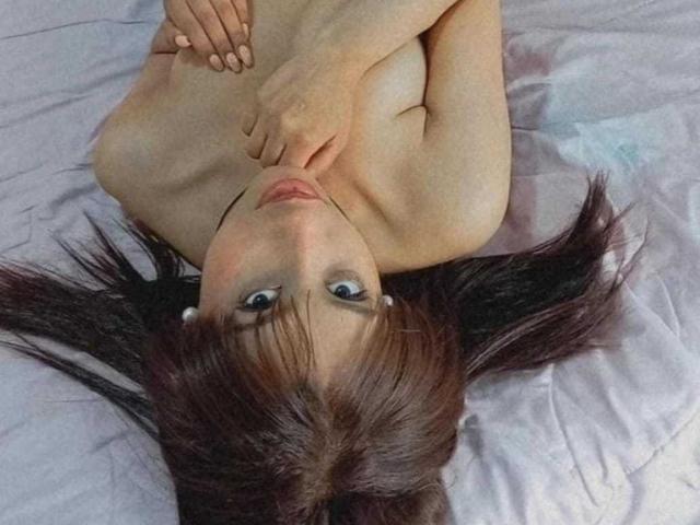 VanessaLove is lying nude on a bed, her hands covering her small tits, with her dark brown hair spread out and her blue eyes looking up at the camera.