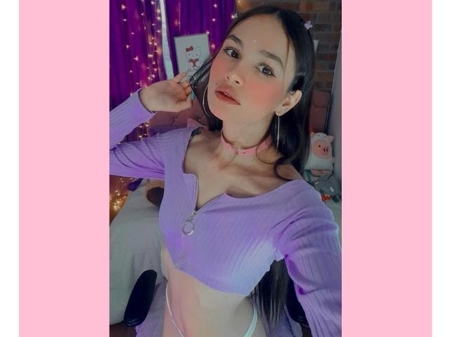 LiliiMoore with long brown hair, wearing a purple ribbed crop top, pink choker, and pink earphones, showing her small tits and flat ass, standing in a pink-lit room.