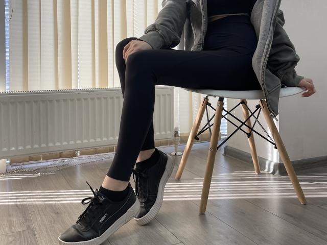 MiaMoone's legs in black leggings, wearing a gray jacket and black Adidas sneakers, sitting on a white chair with wooden legs in a sunlit room.