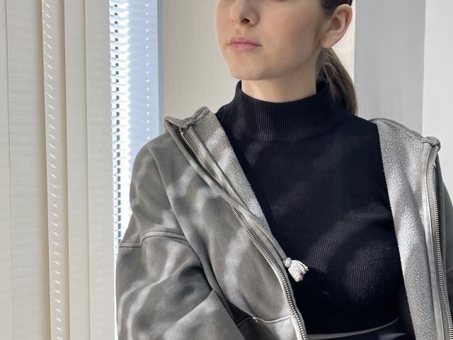 MiaMoone indossa una giacca argentata con zip e un maglione nero a collo alto, che lascia intravedere i suoi seni attraverso il tessuto, con i capelli castano chiaro raccolti in una coda di cavallo, in piedi vicino a una finestra..