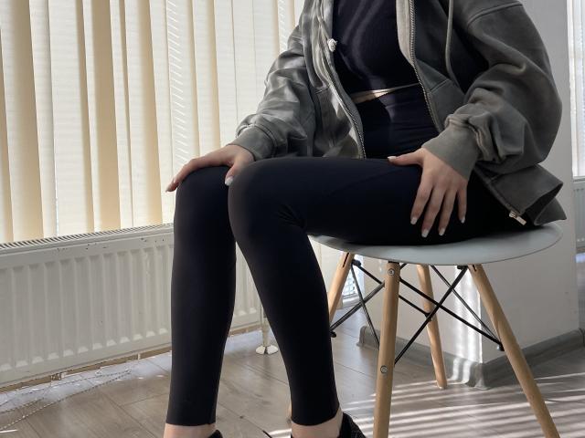 MiaMoone sits on a chair, wearing a gray jacket, black top, and black leggings, with hands on her knees, showing her ass and legs, in a sunlit room.