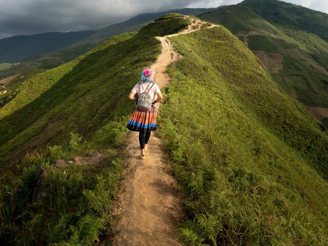 SuperbissimaTroiana indossa un abito colorato, un cappello blu e uno zaino, e sta percorrendo a piedi un sentiero verde su una montagna, con un percorso sterrato, indossando pantaloni neri, vista da dietro..
