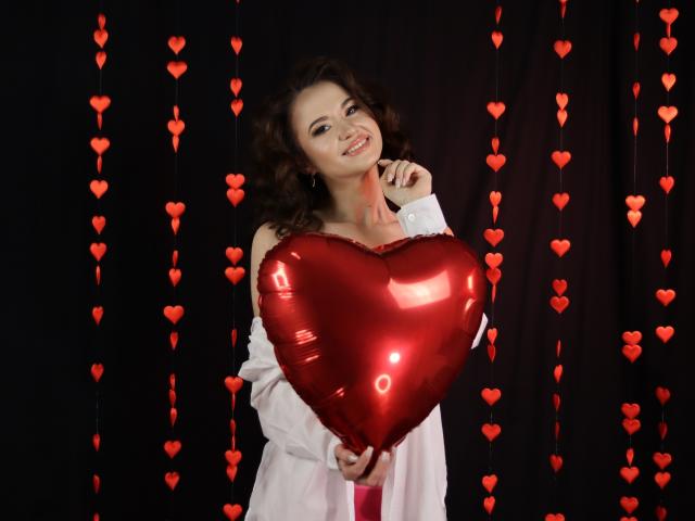 KaraCriegs, con cabello castaño rizado, sonriendo, sosteniendo un globo rojo brillante con forma de corazón, vestida con una camisa blanca, rodeada de decoraciones rojas con forma de corazón colgando..