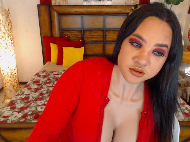 MistressBebe, con cabello negro largo, sombra de ojos roja y lápiz labial naranja, vestida con un cárdigan rojo, mostrando sus grandes senos, mirando a la cámara en un dormitorio..