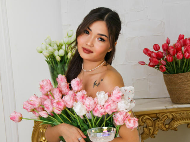 TeyaFaber with pink and white tulips, holding a glass, has dark hair, a tattoo on her chest, and wears a necklace, looking at the camera with a soft smile.