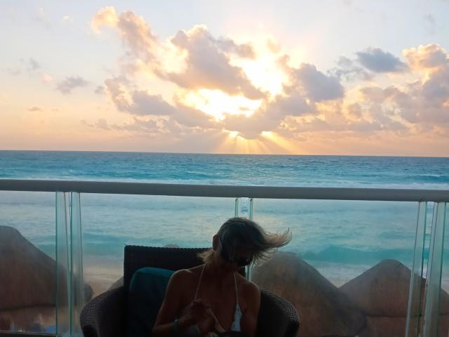 Sunset over ocean, CoupleMature with blonde hair in a white bikini, small tits, sitting on a black chair, glass balcony, blue sea, golden sunlight, scattered clouds.