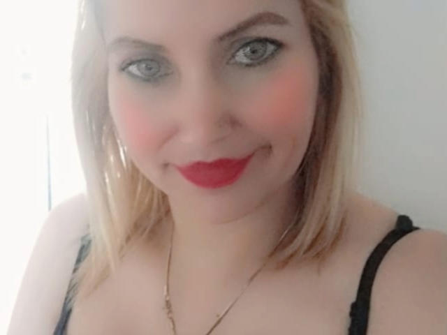 GiuliaAddams with blonde hair, blue eyes, and red lipstick, smiling at the camera, wearing black straps and a gold necklace, showing her tits.