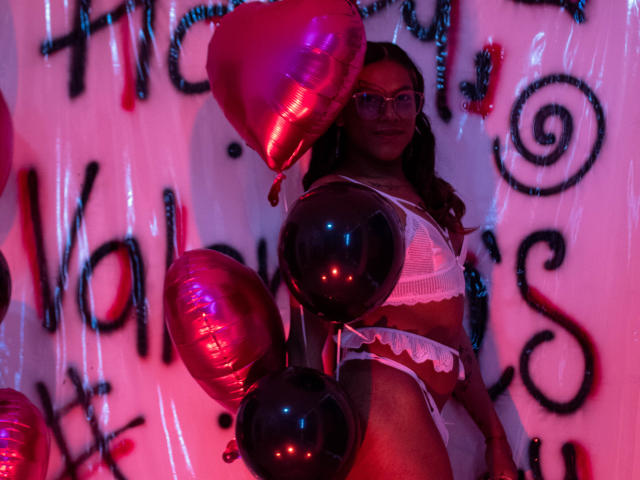 KimmySolange con globos rosas y negros, vistiendo lencería blanca de encaje, mostrando sus pechos y glúteos, en una habitación iluminada en rosa, usando gafas, con cabello largo y castaño..