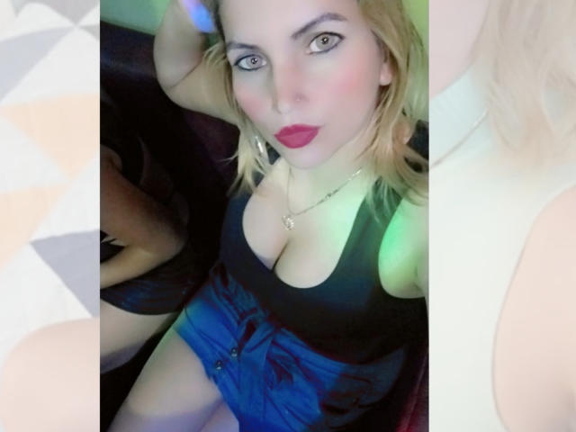  GiuliaAddams has blonde hair, blue eyes, red lipstick, and is wearing a black top with a deep cleavage, showing her big tits, and blue shorts.