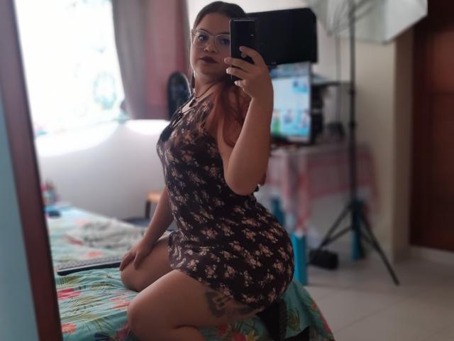 NymeriaStark mit Brille, braunes Blumenkleid, sitzend auf dem Bett, Hintern sichtbar, Handy haltend, Spiegel-Selfie, Schlafzimmer, hellbraune Haut, dunkelbraunes Haar, Tattoos am Oberschenkel..