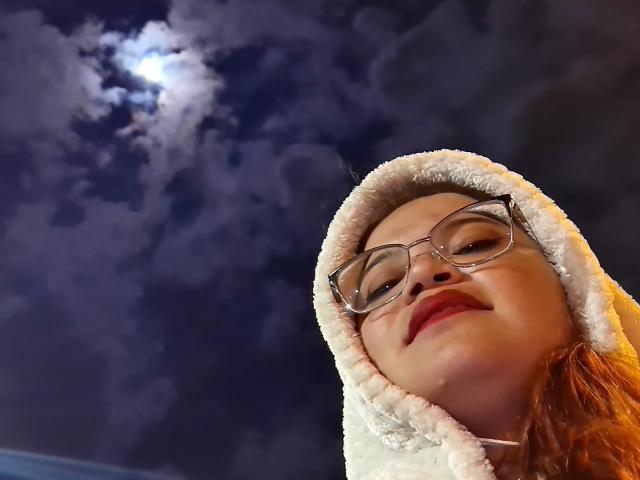 NymeriaStark mit rotem Lippenstift, Brille und einer weißen, flauschigen Kapuze, die in einen mondbeschienenen Nachthimmel mit einem bewölkten, dunkelblauen Hintergrund blickt. Ihr Gesicht ist leicht geneigt und sie lächelt..