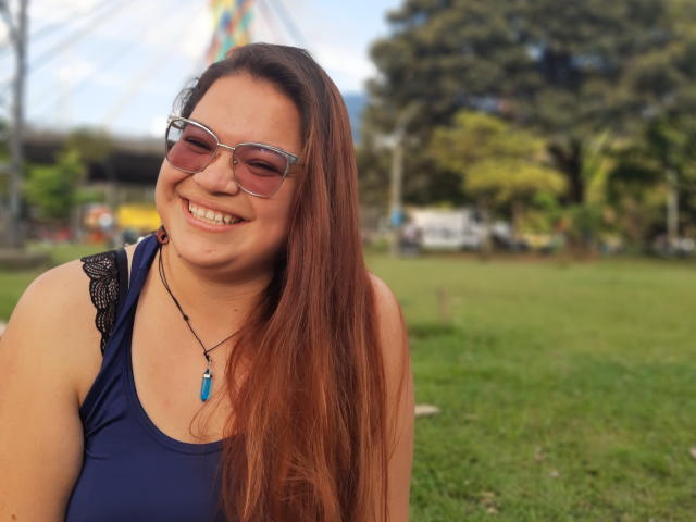 NymeriaStark with long red hair, wearing blue tank top with black lace, smiling, glasses, blue pendant, sitting on grass, trees in background, sunny day, happy face.