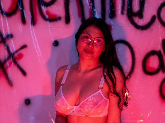 IsabellaPrincess with long black hair, wearing clear glasses and pink lace bra, showcasing her tits under pink and red lighting. Her smile is relaxed, and she's centered.