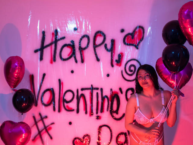 IsabellaPrincess with glasses, white lace bra, and pink sheer top, holding black and pink heart balloons, pink and red lighting, looking at camera. Boobs, glasses, and balloons visible.