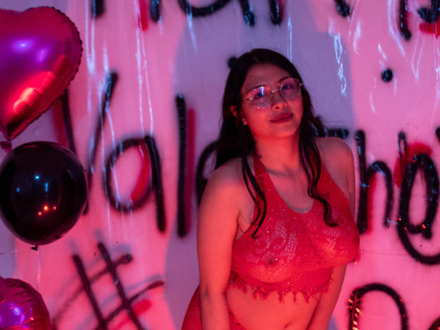 IsabellaPrincess with glasses, red lace top, showing her tits, dark hair, pinkish-red lighting, standing with red and black balloons, looking slightly upward.