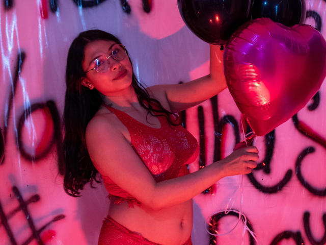 IsabellaPrincess with long black hair, glasses, red sequined bra and panties, holding pink heart-shaped balloons, looking cute and sexy in pink and red lighting.
