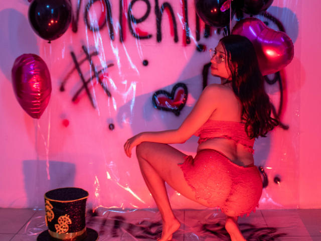 IsabellaPrincess with long brown hair, squatting, wearing pink, sparkly crop top and skirt, exposing her round ass, surrounded by pink and purple balloons, and a black bucket.