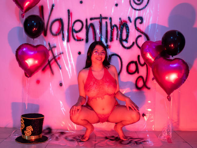 IsabellaPrincess squatting in pink lace lingerie, showing her big tits and pussy, surrounded by red heart balloons, and a black top hat on a tiled floor.