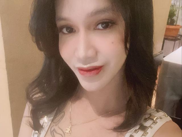 CumEaterJanna with pale skin, dark wavy hair, red lipstick, subtle makeup, and a gold necklace, smiling softly at the camera. Boobs are partially visible under her dress.