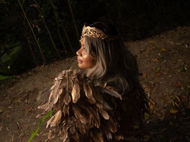AlyssaParker holding a red and gold flame-like object in front of a large, dark red and black coat of arms. The coat of arms is centered and detailed.