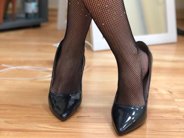 Stefanni's legs in black fishnet stockings and shiny black heels, standing on a wooden floor with a white cable visible.
