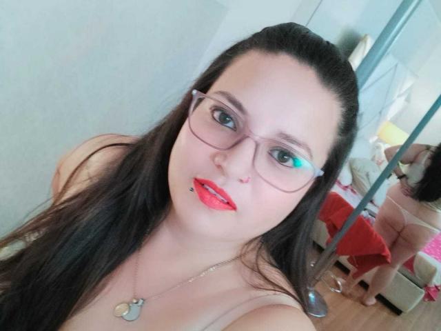 SkayLaraWalker, mit Brille, rotem Lippenstift und einem Nasenring, trägt eine Halskette, hat langes, schwarzes Haar und zeigt auf einem Selfie ihre nackten Brüste..