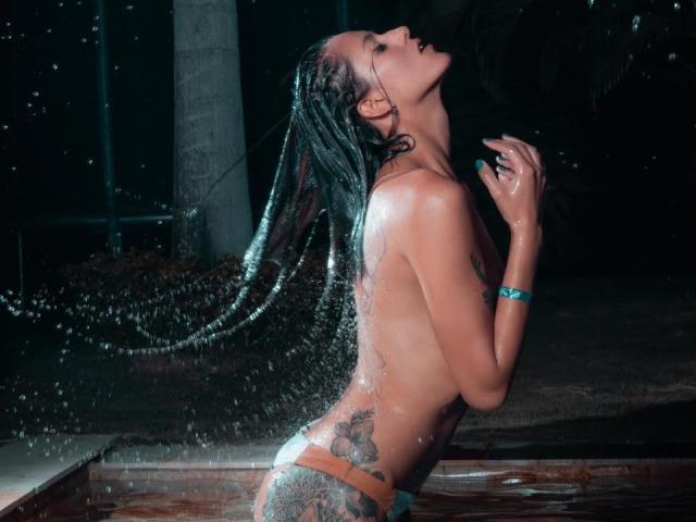VioletteSourire mit nassen, schwarzen Haaren, oben ohne, im Wasser liegend, wobei Tattoos an Seite und Rücken sichtbar sind, Hände nach oben, Wassertropfen, dunkler Hintergrund, blaues Armband, Kopf nach oben geneigt..