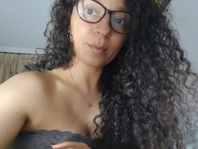 Angelonix with curly black hair, wearing glasses and a lacy black top, exposing her small tits, a delicate necklace, and a subtle smile, in a casual selfie.