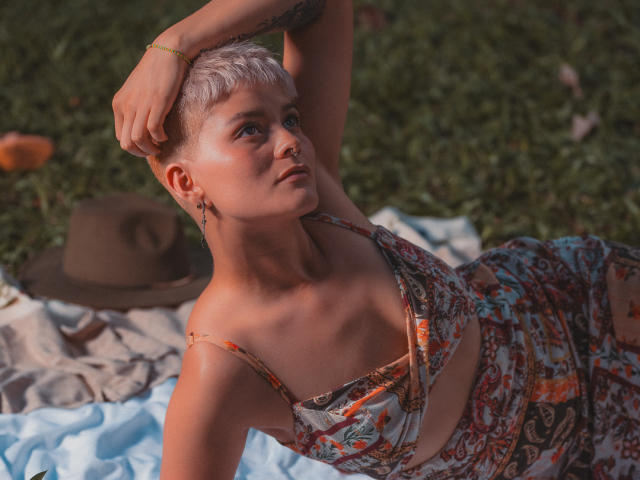 AlexFisher with short, bleach-blonde hair, wearing a patterned dress, sits on a blanket, arm raised, exposing a tattoo, with a brown hat in the background.