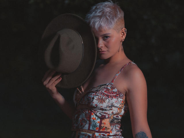 AlexFisher with short white hair, holding a brown hat, wearing a colorful patterned dress, has a nose ring and tattoo, dark background, looking at camera with serious expression.