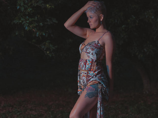 AlexFisher, met kort grijs haar, draagt een bloemenprintjurk die doorschijnend is en kleine borsten en een kleurrijke tatoeage op haar dij laat zien. Ze staat in een donker bos, met haar hand in haar haar, en kijkt naar beneden..