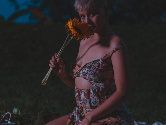 AlexFisher with short gray hair, holding a sunflower, wearing a floral off-shoulder top and matching skirt, sitting in dark, showing small tits, and a cute ass.