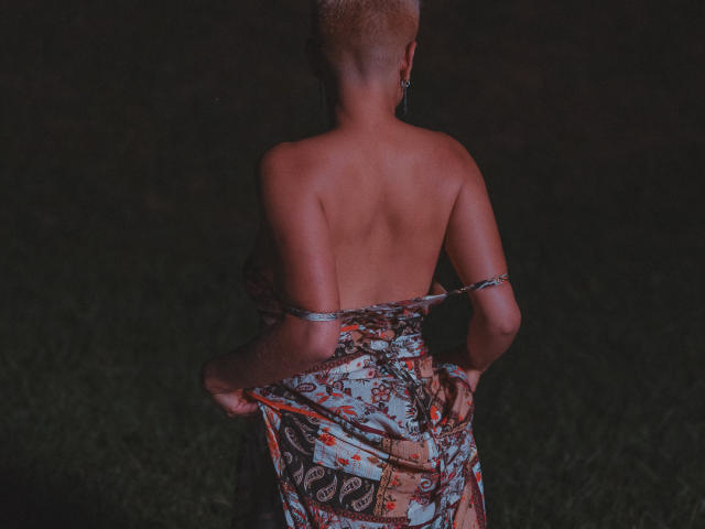 AlexFisher with short blonde hair, bare back, and a colorful patterned dress pulled down, exposing her ass, in a dimly lit, grassy setting.