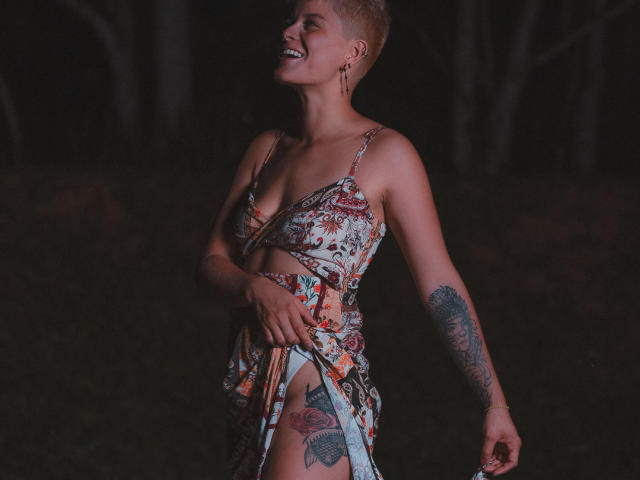 AlexFisher with short blonde hair, smiling, wearing a floral dress, revealing tattoos on her right thigh and left arm, standing in a dark forest at night.