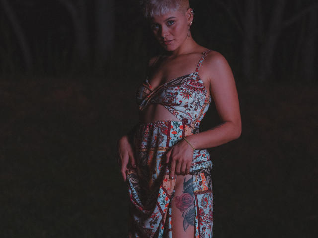 AlexFisher with short blonde hair, wearing a floral dress, exposing her tits and a large tattoo on her thigh, standing in dark, moody lighting.
