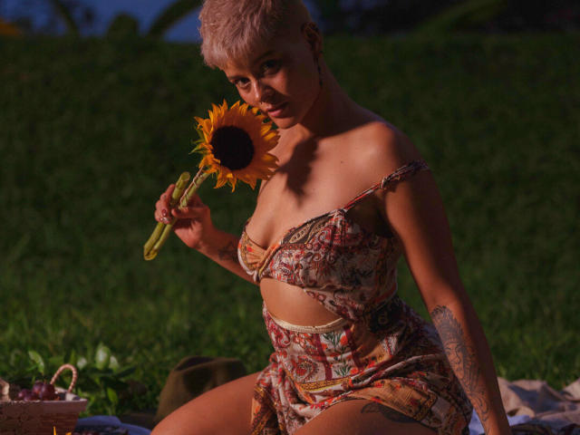 AlexFisher, met kort roze haar, die een zonnebloem vasthoudt, gekleed in een kleurrijke, gemêleerde outfit met een open schouder en een blootliggende buik, zittend op het gras, met een tatoeage zichtbaar op haar rechterarm..