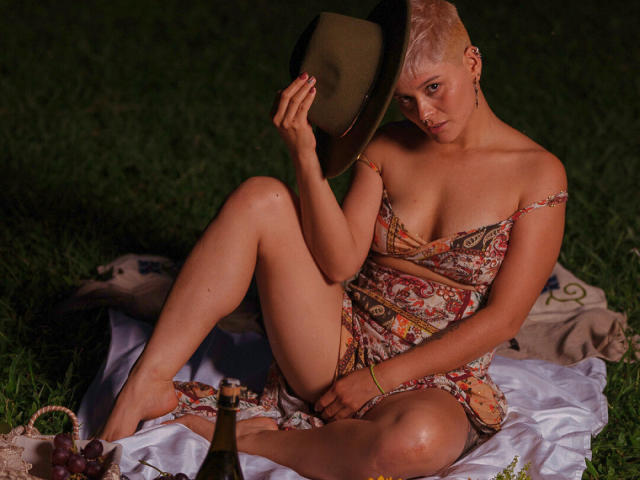 AlexFisher, sitting on grass, wearing a patterned dress, holding a hat, showing her tits, with a bottle and grapes on a white blanket, night, short blonde hair.