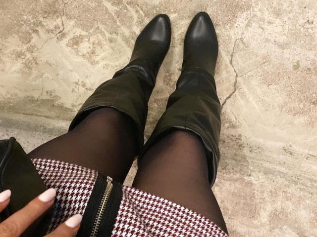 ArianaCortes' legs wearing black leather boots, houndstooth skirt with a zipper, and black tights, standing on a textured concrete floor, showing a hand with pink nails.