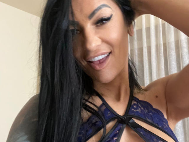 AndraD has long black hair, dark eyebrows, and is smiling with her tongue out. She's wearing a lace blue top with black straps, exposing her tits.