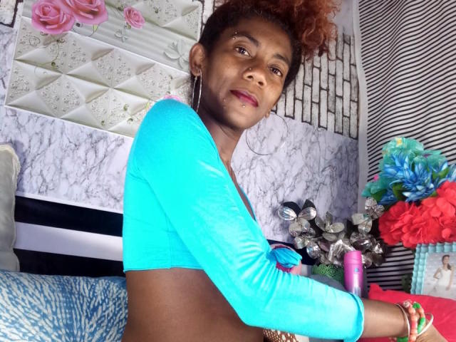 GinaSensual69 met bruine huid, krullend rood haar, draagt een blauwe top met lange mouwen, toont kleine borsten, houdt een roze vibrator vast, heeft een rode neusring en kleurrijke armbanden, en kijkt naar de camera..