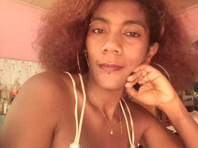GinaSensual69, con i capelli rossi ricci, la pelle marrone, gli orecchini a cerchio, la collana d'oro e il piercing al labbro, sorride alla fotocamera, mostrando il seno con una canotta bianca a spalline..