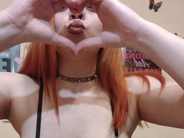 SpicyGrace mit orangefarbenem Haar, die mit ihren Händen ein Herz formt, die Lippen zusammenpresst und ein schwarzes Top mit Trägern und eine Halskette mit Nieten trägt. Ihre Brüste sind sichtbar..
