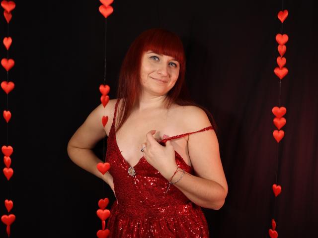 NinaFlawlees with red hair, wearing a sparkly red dress, pulling down a strap, showing a small nipple, surrounded by red heart decorations, smiling.