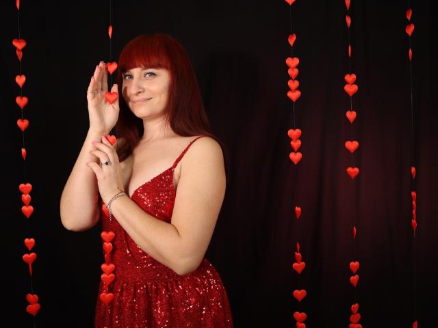 NinaFlawlees with red hair, wearing a sparkly red dress, holding a red heart, smiling, standing against a black background with hanging red hearts. Boobs visible through dress.