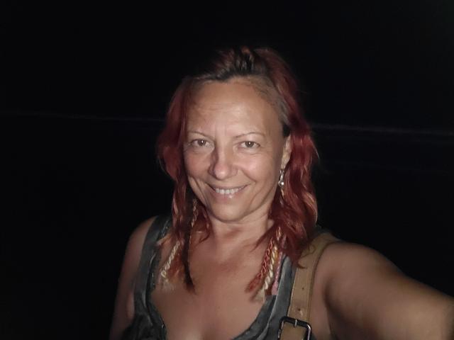 BarbaraBowie with red, wavy hair, smiling, wearing a gray tank top and brown straps, showing small tits, in a dark, nighttime setting, taking a selfie.
