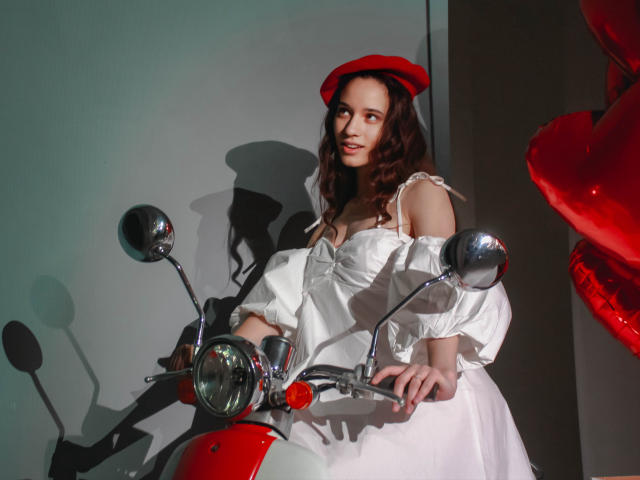 StacieLove, avec ses cheveux bruns ondulés, portant une casquette rouge et une robe blanche à épaules dénudées, est appuyée contre une moto rouge et blanche, souriante, avec des rétroviseurs brillants devant elle..