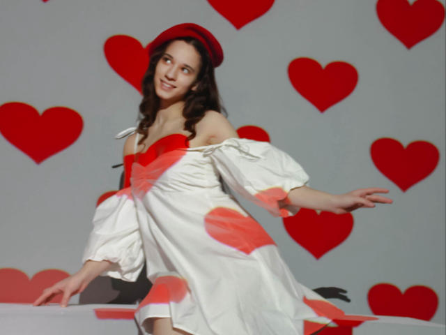 StacieLove has long brown hair, wearing a red beret, off-shoulder white dress with red hearts, and is smiling with arms slightly raised, standing against a grey wall.