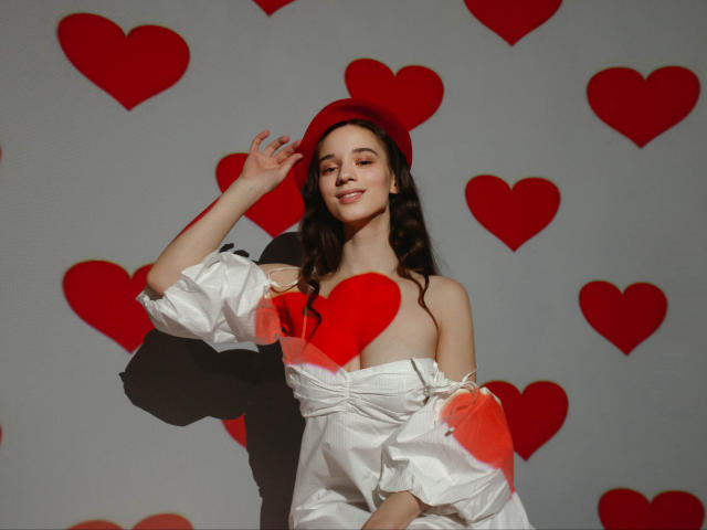 StacieLove with long dark hair, wearing a red beret and white off-shoulder dress with red heart patches, smiling, hand on beret, red heart decals in background.