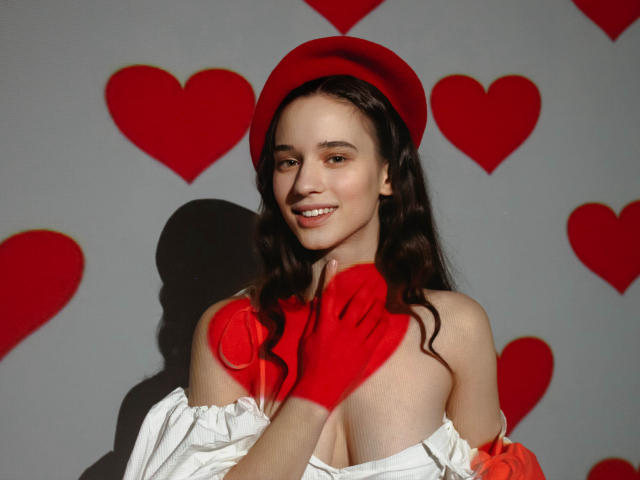 StacieLove with red beret, red gloves, and white off-shoulder top, smiling at camera, showing small tits, wavy brown hair, and cute face, against grey wall with red hearts.